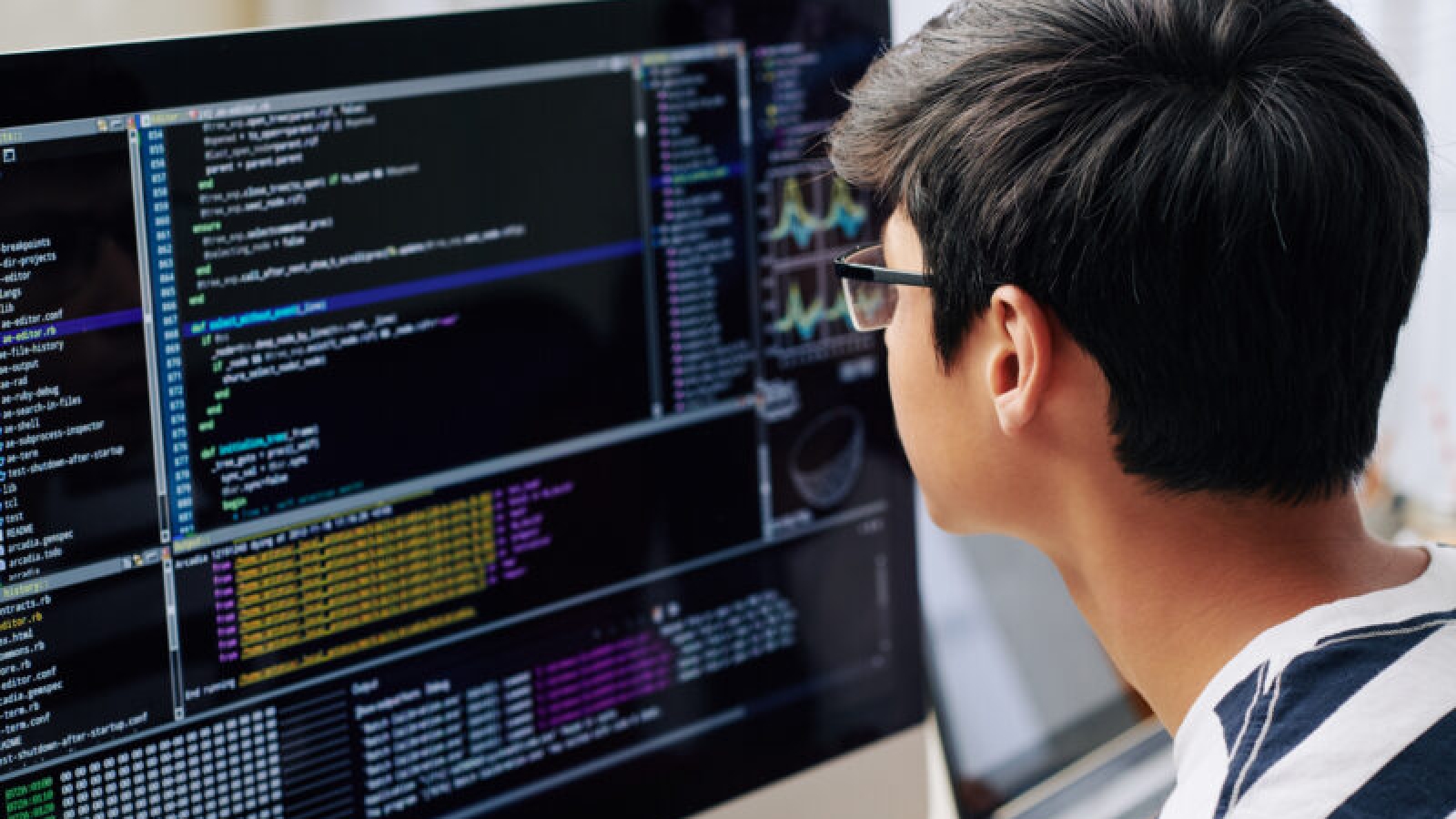 Smart teenage boy in glasses checking programming code on computer screen when sitting at his desk